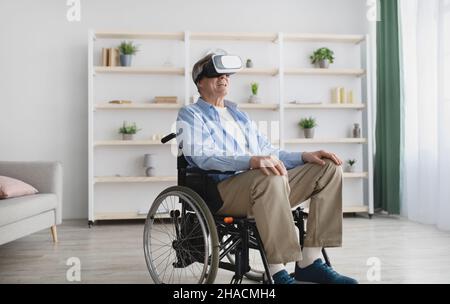 Happy senior man in wheelchair wearing VR glasses, playing online video game in cyberspace at home Stock Photo