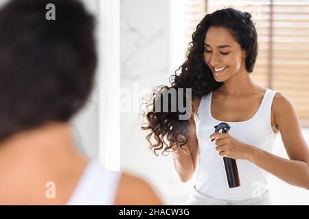 Haircare. Beautiful Young Lady Applying Hair Spray, Standing Near Mirror In Bathroom Stock Photo