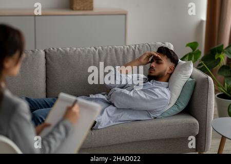 Young arab male patient lying on couch at psychologist's office, having session with counselor, guy feeling depressed Stock Photo
