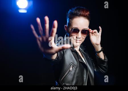 Stop, i don't want to see. Portrait blocking woman with short hair and makeup in black leather jacket in casual style, standing with a stop gesture in sunglasses. Black background with blue light Stock Photo