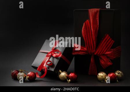 Composition of gifts of different shapes wrapped in black wrapping paper with a red ribbon and a bow on a black background. Side view. Copy space. Stock Photo
