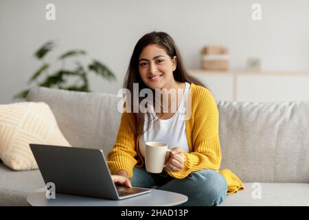 Portrait of happy arab female freelancer drinking coffee and using laptop, working online from home, copy space Stock Photo