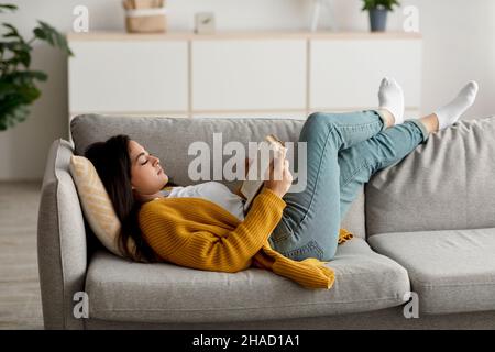 Cozy stay at home activities. Young arab woman reading her favorite book, lying on sofa in living room interior Stock Photo