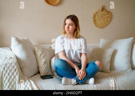 Stressed young woman sitting on sofa in casual clothes, worrying about problems alone at home, afraid of difficulties, looking away. Nervous frustrated lady sitting on the couch feeling envy. Stock Photo