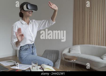 Joyful young business woman sitting on top of desk at home wearing VR glasses Stock Photo