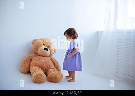 Little girl in lavender purple dress stands next to a big teddy bear Stock Photo