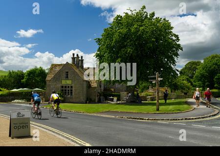 2 cyclists & walkers passing quaint attractive cottage tea rooms cafe in scenic sunny rural village - B6160 Bolton Abbey, Yorkshire Dales, England UK. Stock Photo