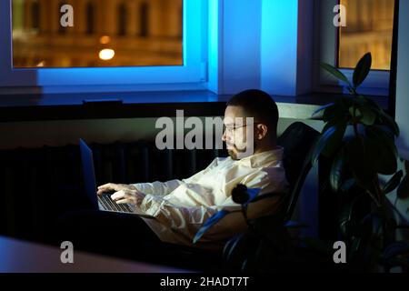 Tired programmer in dark office work on laptop. Workaholic businessman typing on computer at night Stock Photo