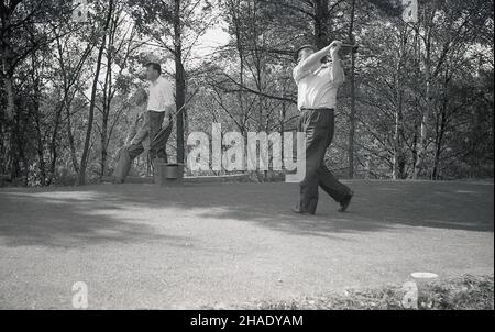 1960s, historical, on a woodland inland golf course, a male golfer hitting off a tee, watched by his playing partner and caddie, Engand, UK. Stock Photo
