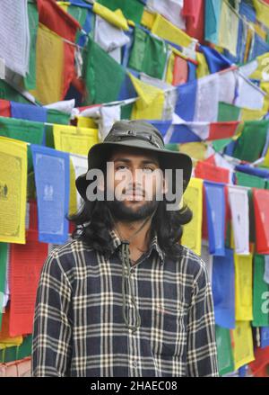 A bearded and long haired male tourist  looking at camera while standing against Buddhist prayer flags Stock Photo