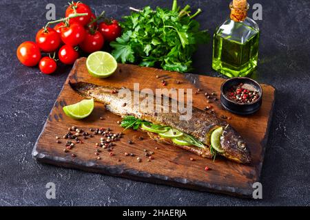 whole trout fish baked in an oven with herbs and spices, served on a wooden cutting board with lime slices and parsley Stock Photo
