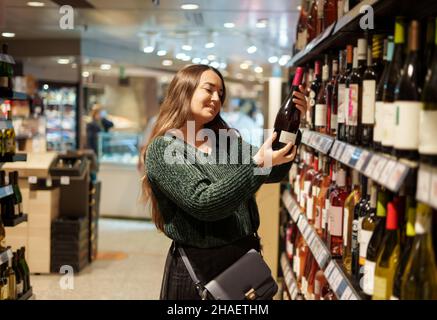 Happy young woman choosing favorite red wine standing by shelves with different types of alcoholic beverages in grocery store, looking forward to celebrate New Year with family in cozy home atmosphere Stock Photo