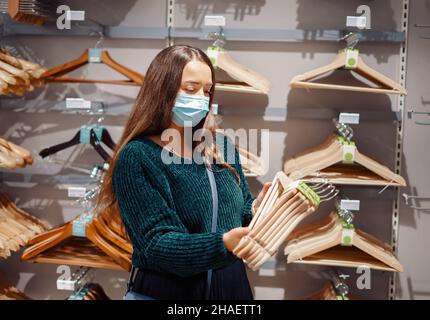 Young woman in face mask buying household products, choosing goods in store everything for house, holding set of wooden hangers in hands, doing shopping in mega Mall during covid-19 pandemic Stock Photo