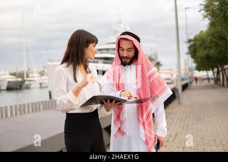 Smiling saleswoman explaining contract documents to arab businessman in traditional clothes while standing in port, attractive europian businesswoman discussing deal conditions with saudi man client Stock Photo