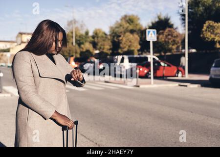 Beautiful African American woman holding suitcase and looking at wristwatch. Holiday and travel concept Stock Photo
