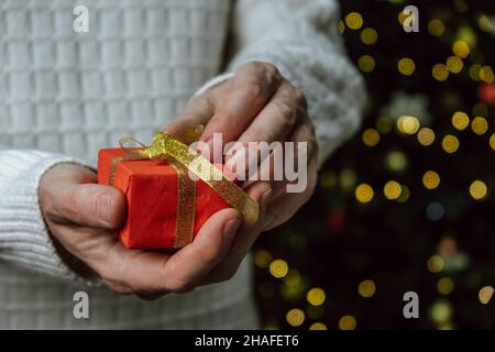 Christmas New Year card. A man in a white sweater holds an elegant red gift box on festive Christmas background. Pleasant holiday surprises and gifts. Stock Photo