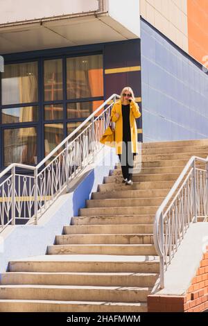 Elegant modern Woman with blond hair in a bright yellow coat and sunglasses walks along a city street. Urban style Stock Photo