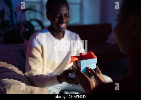 African man receives unexpected present from wife at home, woman giving gift box to happy boyfriend Stock Photo