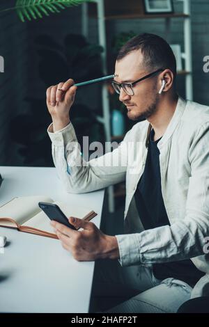 Portrait of thoughtful bearded man wearing eyeglasses using cellphone while working at home. Stock Photo