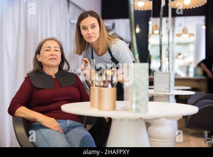 Young woman professional makeup artist working in beauty studio with positive elderly female client, applying tonal foundation on face. Stock Photo