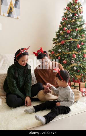 Young parents playing with little son when sitting on the floor at home next to Christmas tree Stock Photo