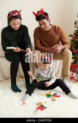 Young parent wearing reindeer antlers headbands when looking at their child playing with toys on Christmas eve Stock Photo