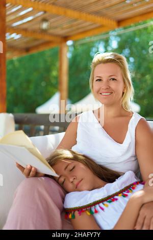 Caucasian middle aged woman reads a book while her daughter takes a nap in her lap on a summer garden. Sunny day. Focus on mothers face. Shallow depth of field. Stock Photo