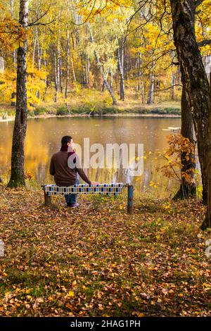 Man in a sweater and scarf sits on a bench by a pond in the fall and enjoys the view. Stock Photo