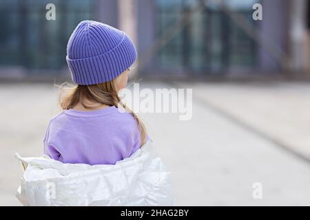 Cute little caucasian girl eight years old with blonde hair standing from behind outdoor. Kid wearing stylish shirt and knitted hat violet color Stock Photo