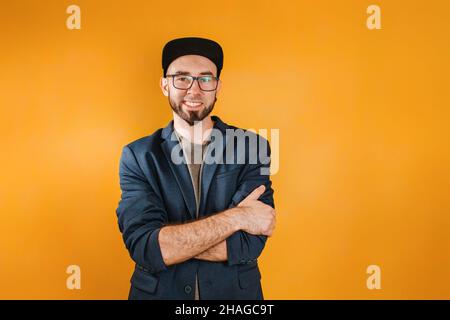 The concept of success and business. A man in a jacket and cap, with a beard and glasses, put his arms around his shoulders and smiled. Orange backgro Stock Photo