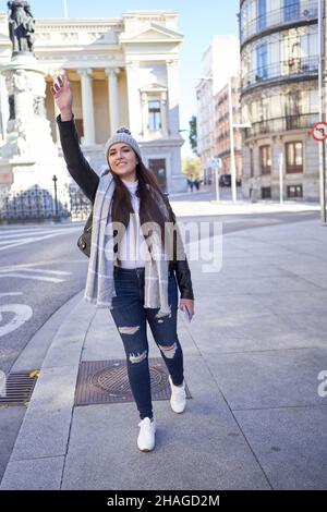 Full body of positive young female millennial with long dark hair in casual clothes and hat standing on sidewalk with raised arm while catching taxi, on city street Stock Photo