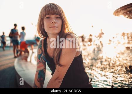Portrait of tanned young woman with a tattoo on her arm is sitting by the fountain. Sunset in the background. Stock Photo