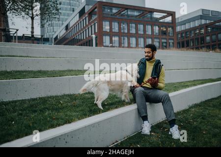 Happy young man sitting on grass with his dog outdoors in town. Stock Photo