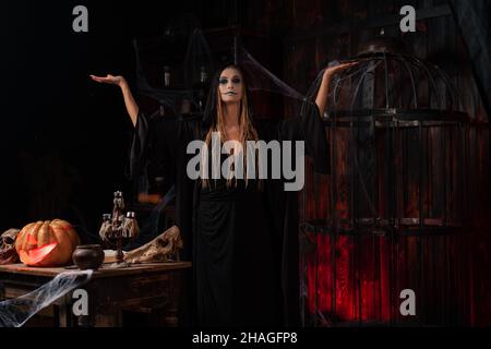 Halloween concept. Witch dressed black hood with dreadlocks standing dark dungeon room rise hand for conjuring magic spell. Female necromancer wizard Stock Photo