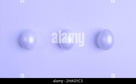 Three glossy Easter eggs are lying in row. Creative minimal festive Easter background. Trendy toning in color of year lavender Very Peri. View from ab Stock Photo
