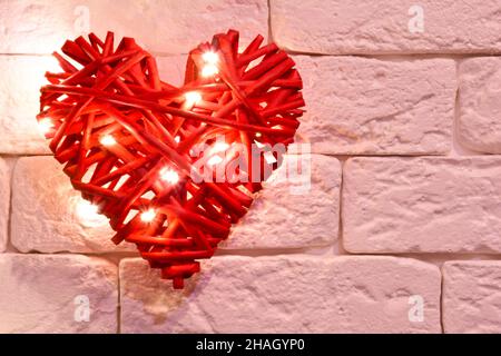 Handmade red wicker decorative heart, illuminated by lights against a light brick wall. There is a place on the right for inscriptions for different h Stock Photo