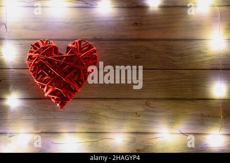 Large red wicker decorative heart on the left side on a wooden background. There are glowing lights around the frame. On the right there is a place fo Stock Photo