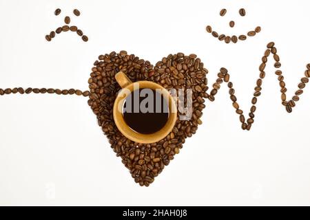 On a white background, there is a cup of coffee, a heart shape is laid out under it from coffee beans and on the sides a cardiogram of a heart pulse Stock Photo