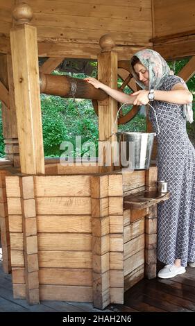 A young girl raises an iron bucket full of water from a wooden old well Stock Photo