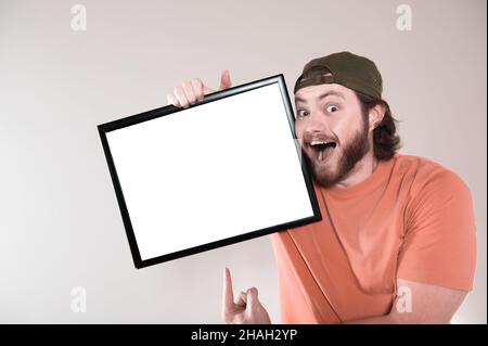 Young bearded man making funny face, holding a blank sign board. Studio shot on gray background. Stock Photo