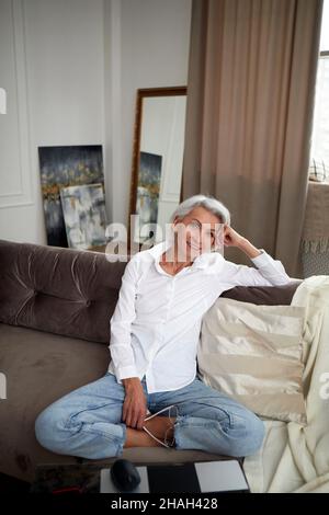 High angle of charming middle aged female with gray hair and in jeans and white shirt sitting with crossed legs on couch at home and smiling at camera Stock Photo