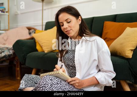 Cropped shot of caucasian woman sits crossed legs, wears home pants and white shirt, makes notes in diary, focused aside, blank copy space against domestic interior Stock Photo
