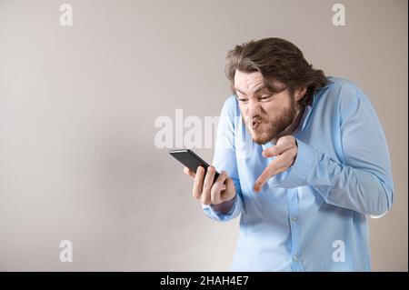 Aggressive young business man in blue shirt shouting into his mobile phone on grey background Stock Photo