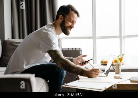 Bearded european man using mobile phone while working with laptop at home Stock Photo