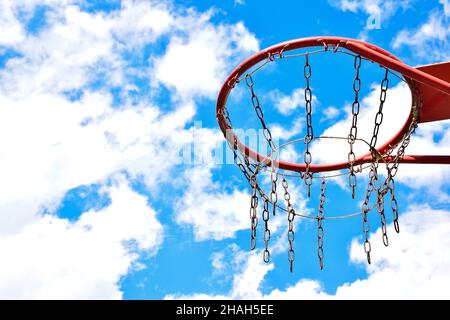 Close-up of a basketball hoop on the right side against a bright blue sky with white clouds. On the left side there is an empty space to insert text Stock Photo