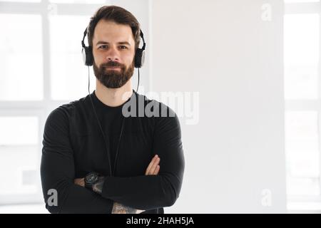 Young smiling brunette bearded white man in sportswear standing indoors listening to music in headphones Stock Photo