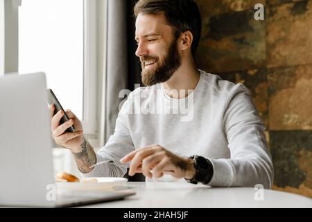 Bearded man smiling and using cellphone while working with laptop indoors Stock Photo