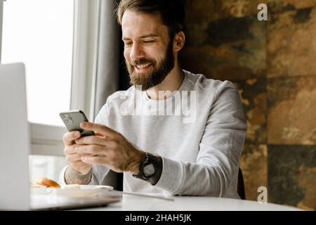 Bearded man smiling and using cellphone while working with laptop indoors Stock Photo