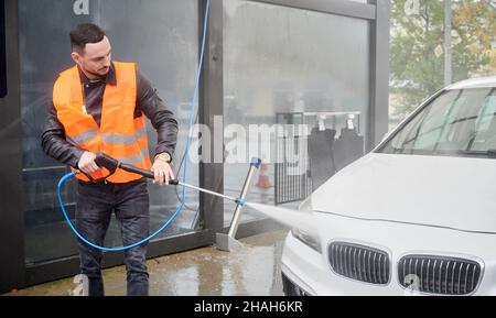Young man washing car on carwash station, wearing orange vest. Handsome worker cleaning white automobile, using high pressure water. Stock Photo