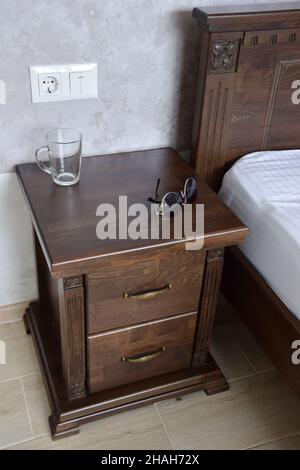 Sunglasses and an empty glass lie on a decorative brown wooden nightstand near the bed. Wall mounted electrical outlet and light switch Stock Photo